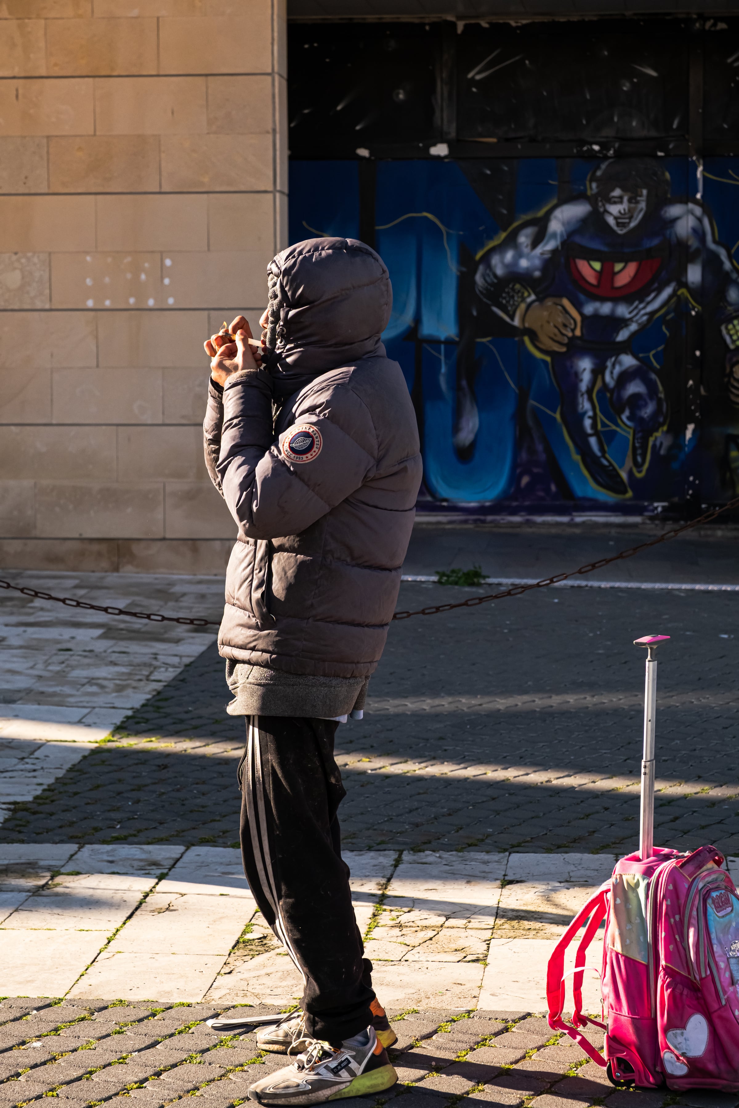 Figure in heavy jacket facing away from camera with superhero graffiti mural and pink child's suitcase