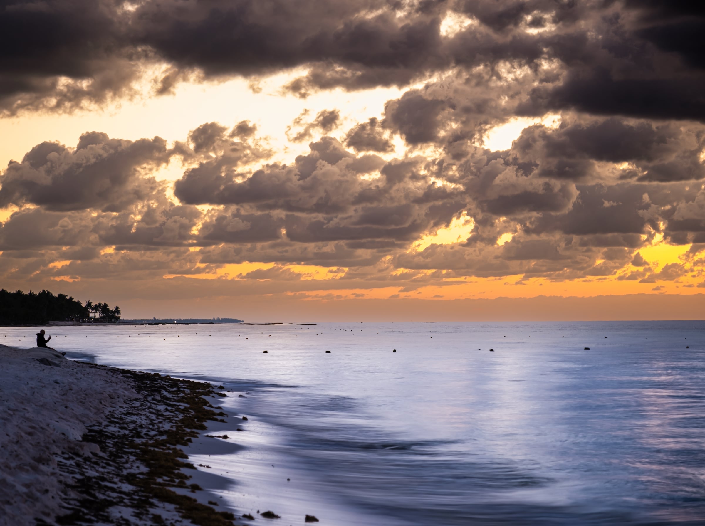 Caribbean beach at dusk with lone silhouette sitting on shoreline and dramatic golden-orange sky