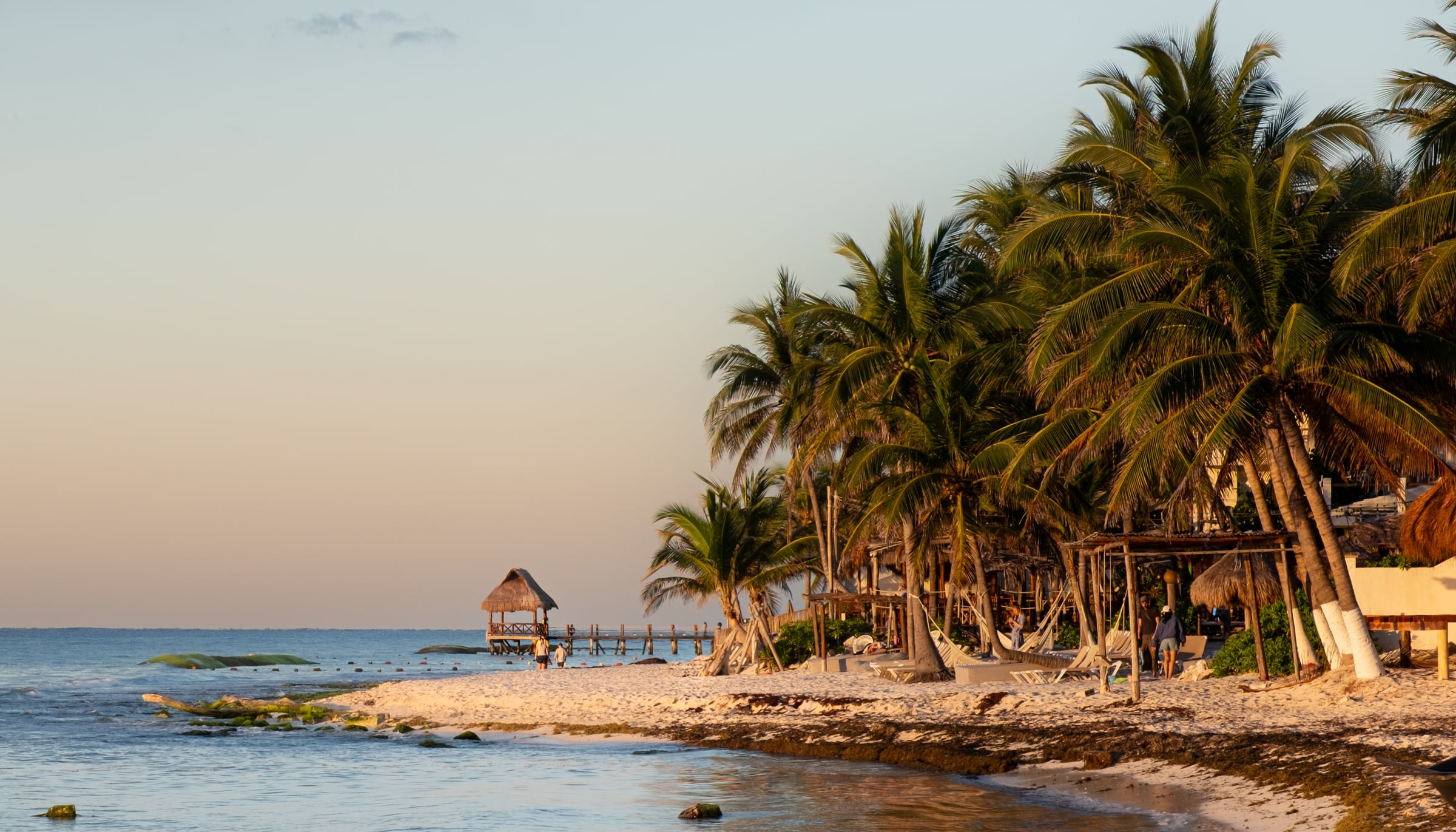 Tropical beach with coconut palm trees, palapa huts and wooden pier at golden hour