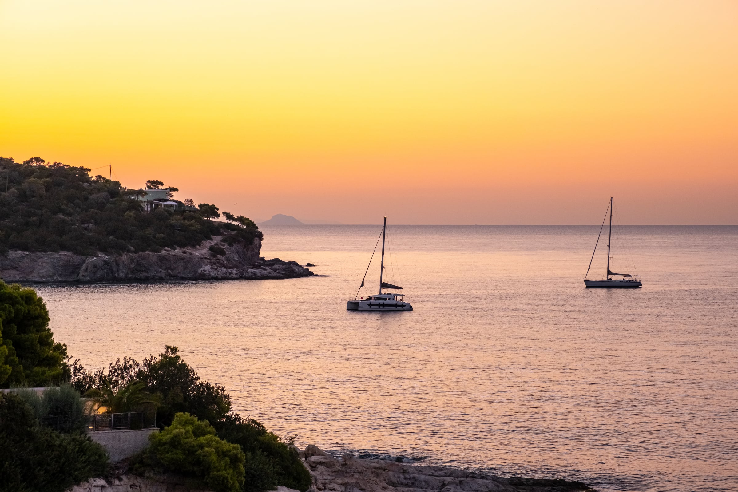 Two sailboats at golden sunset in a calm Mediterranean cove with rocky forested headland