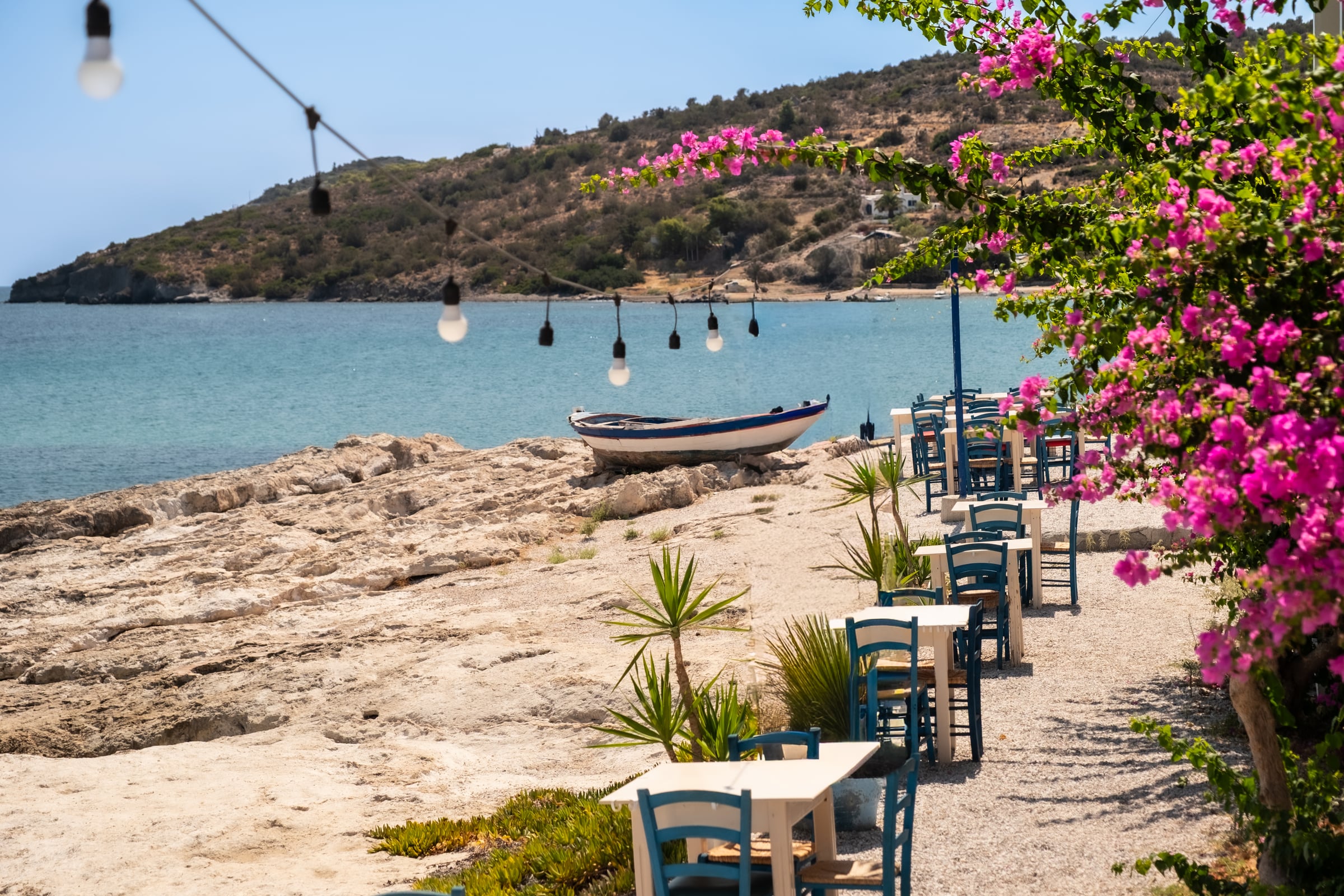 Greek waterfront taverna with blue-white chairs, old fishing boat on rocks, pink bougainvillea, string lights