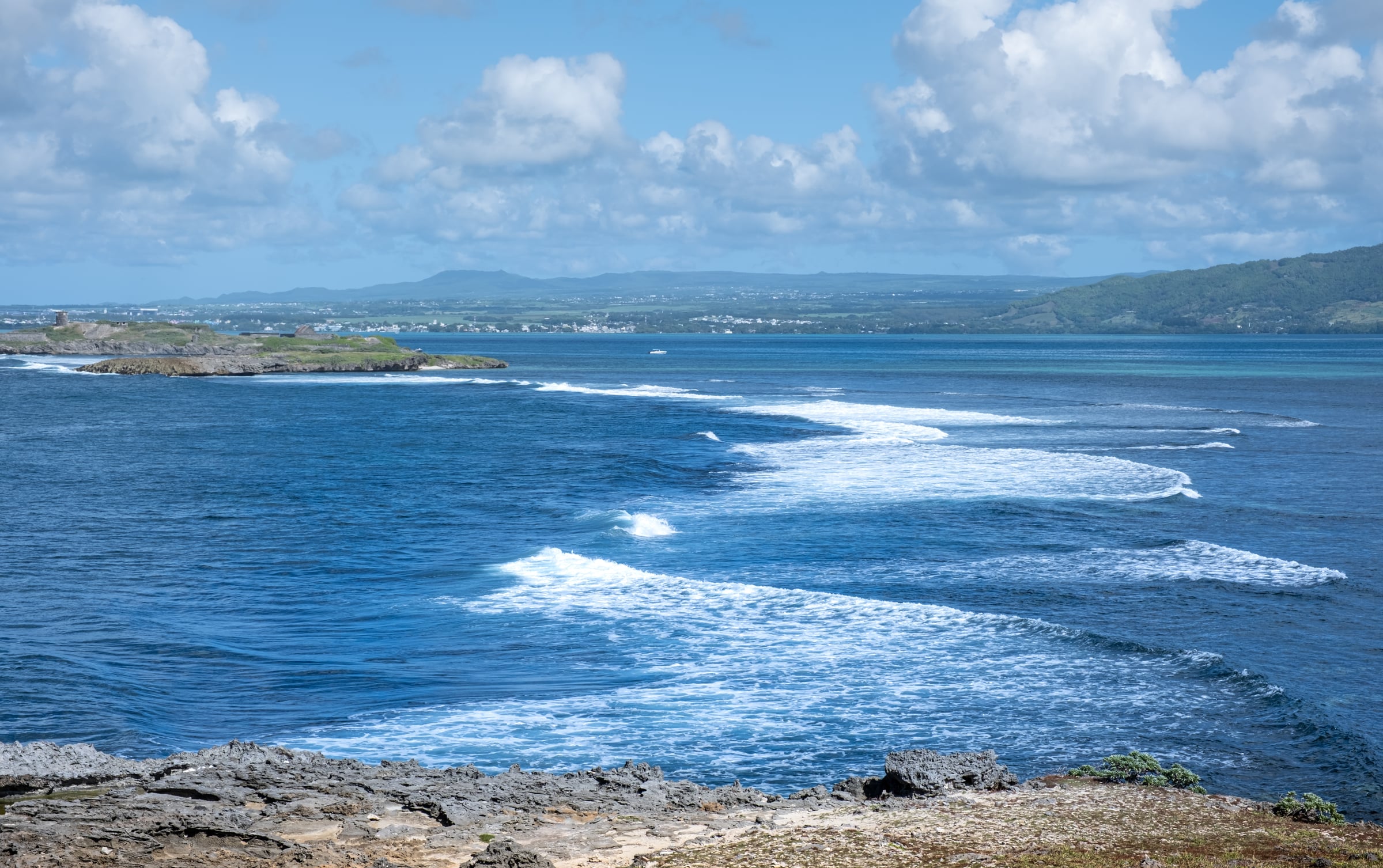 Ocean waves breaking in lines along rocky tropical coastline with deep blue sea and green hills