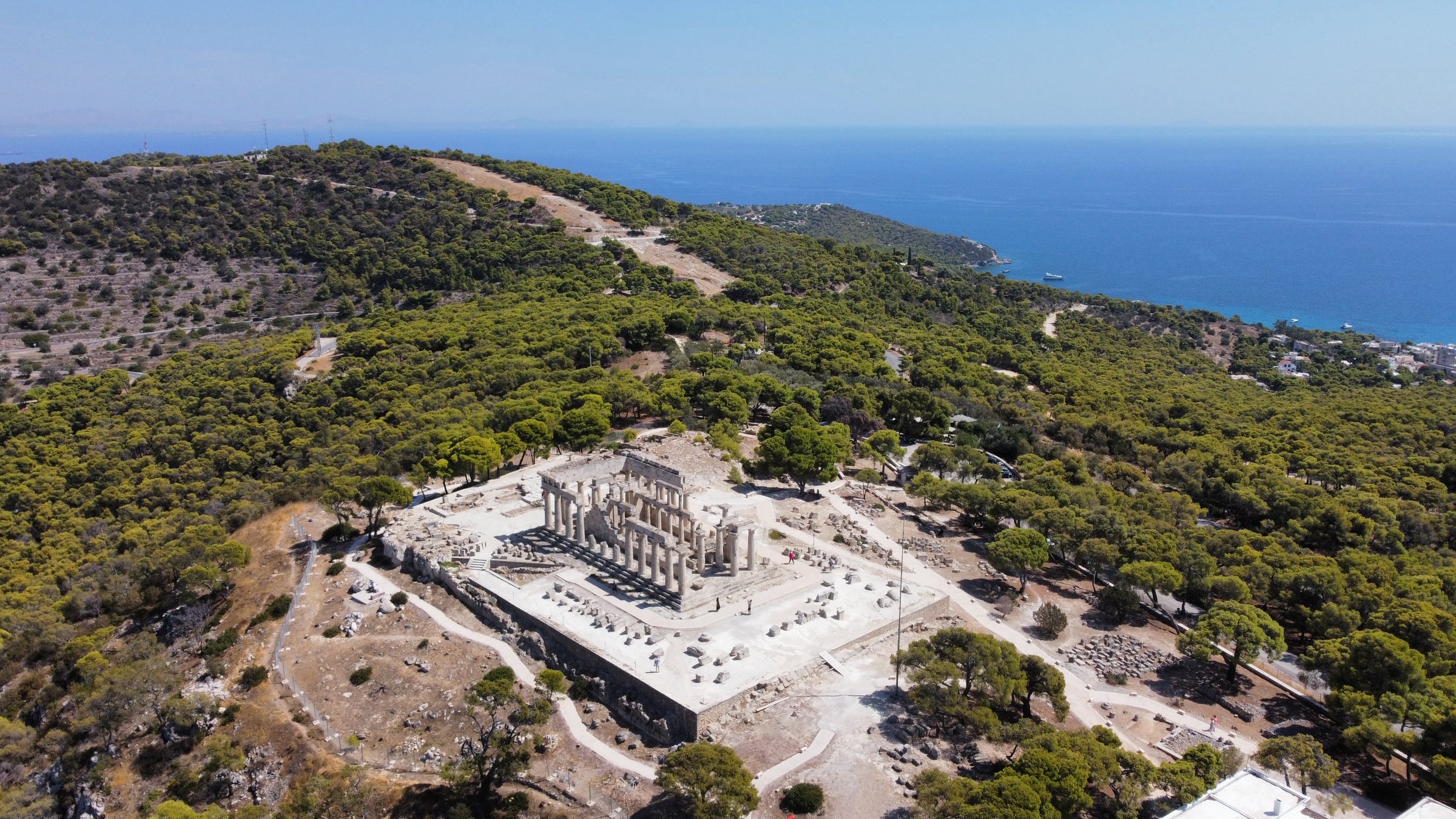 Wider aerial drone view of Temple of Aphaia surrounded by pine forest on Aegina hilltop with Mediterranean sea
